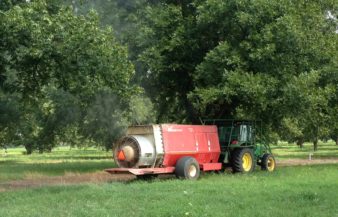 A tractor pulls a sprayer behind it. The sprayer is shooting a fine mist in to the trees behind it.