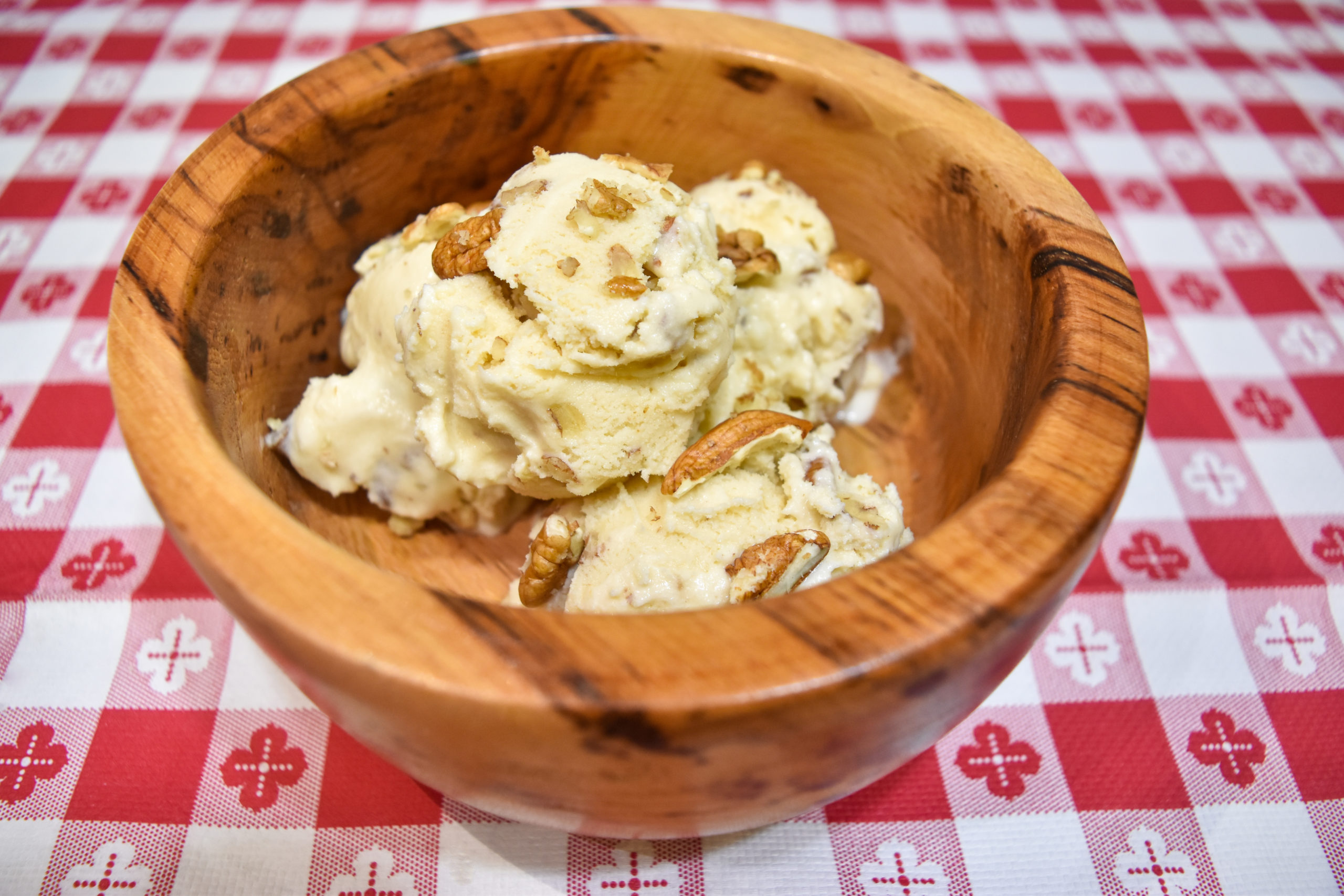 Three scoops of butter pecan ice cream sit in a wooden bowl on a gingham tablecloth.