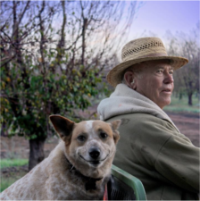 A man wearing a hat sits in his cart with his dog in front of some trees.