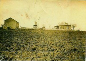 A sepia-toned photo showing a tilled field with a barn and house in the background.