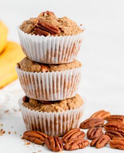 Three Banana Pecan Muffins stacked on top of each other with pecan halves scattered in front and a banana in the background.