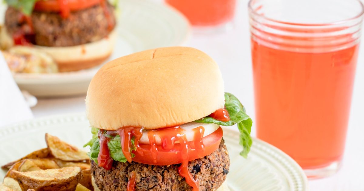 Fully dressed, Black Bean Pecan Burgers sit on plates with homemade fries next to glasses of juice.
