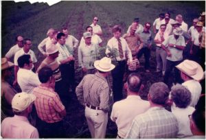 George Ray McEachern stands next to a young pecan tree in the center of a crowd of pecan growers of various ages.