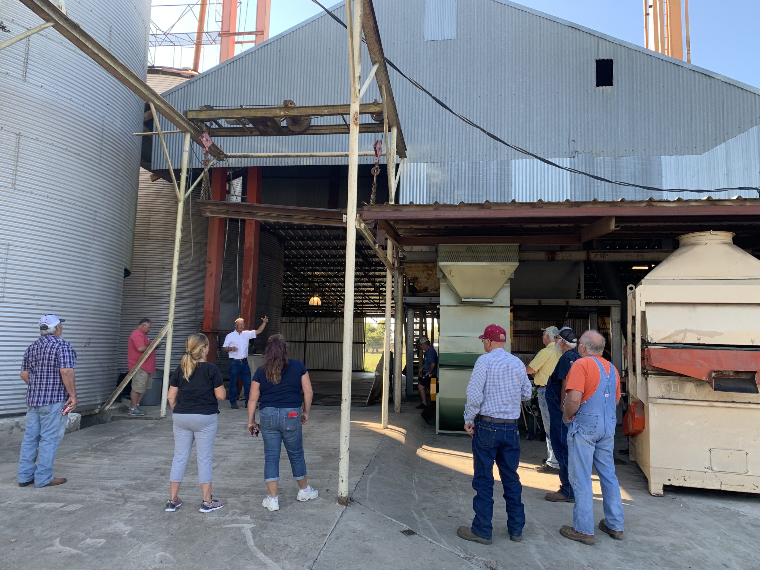 People stand around in small groups listening to a presentation outside of a metal-sided cleaning plant at Tri-Agri Farm Center..