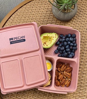A pink plastic bento box with the APC Pecan Powerhouses logo sits open on top of a wicker table. The box holds a handful of blueberries, half an avocado, some shelled pecans, and a hard boiled egg. This box was part of APC marketing activities, which will continue in 2022.