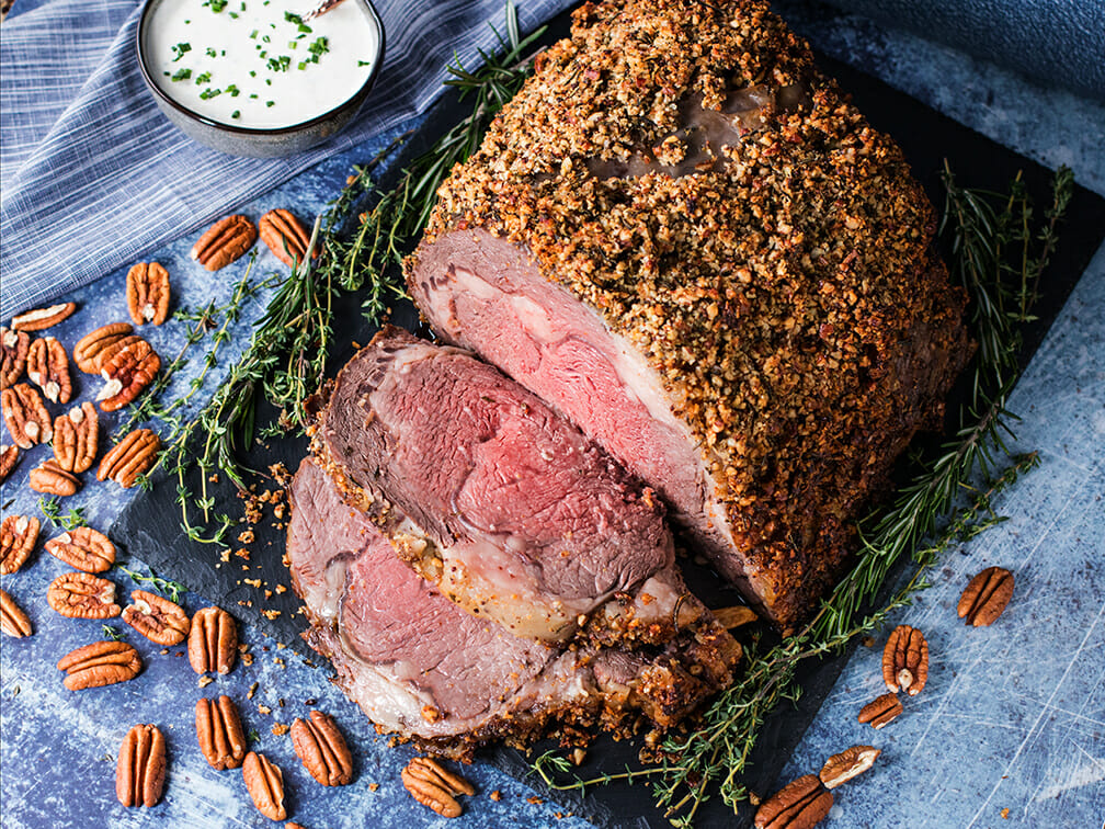 A pecan-crusted prime rib served on a black cutting board surrounded by pecan halves and green herbs. Several pieces of meat have been sliced and lay on display in front of the rib.