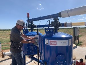 A man in a ball cap, gray shirt, and blue jeans checks a monitor on an bright-blue, double barreled irrigation pump owned by Raptor Ag.