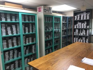 The nut library at the USDA ARS Pecan Breeding and Genetics program has multiple teal shelves filled with little black boxes of nuts and a wooden table. These are some of the nuts that make up the germplasm repository.