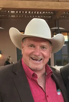 Carl Bissett wears a white cowboy hat and a red polka-dot button down with a brown blazer. He gives a big smile toward the camera.
