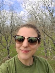 A selfie of a woman wearing sunglesses and a green shirt in a dormant pecan orchard.