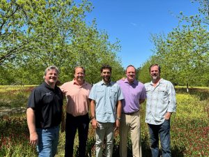 Five men smile at camera in a pecan orchard in early spring.