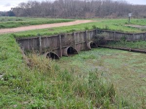 A canal leading underneath a dirt road bridge with four circular gates.