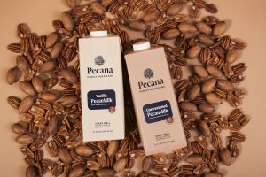Two cartons of Pecana Nutmilk rest on a bed of inshell pecans and pecan kernels against a tan backdrop.